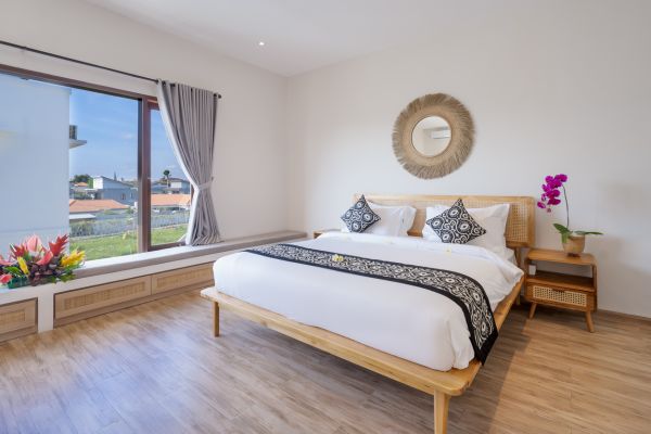 Cozy tropical bedroom with wooden bed, white linens, rattan mirror, purple orchids, and large window overlooking lush green landscape. - Bali Villas