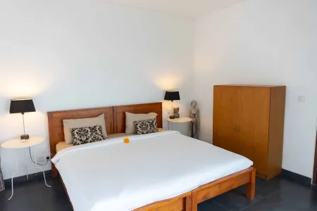 Modern minimalist bedroom with wooden platform bed, white linens, bedside lamps, frangipani flower, and Balinese statue decor.