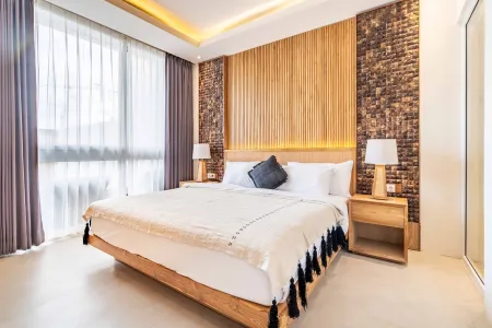 Modern wooden bedroom with floating king bed, white tassel bedding, bedside lamps, and large sheer-curtained windows.