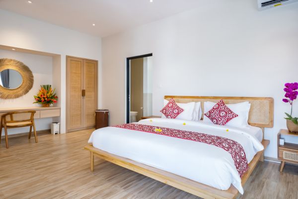 Spacious Balinese villa bedroom featuring a wooden king bed with white linens and red batik runner, rattan accents, orchids, and tropical decor.