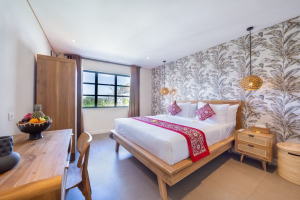 Spacious Balinese-style bedroom with wooden king bed, red runner, tropical leaf wallpaper, pendant lights, and fruit bowl on desk. - Bali Villas