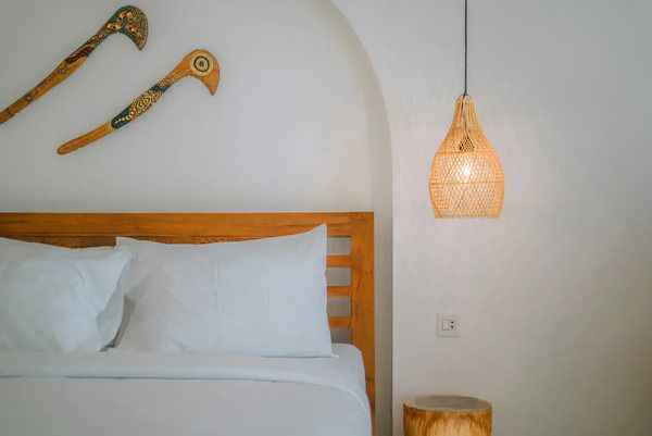 Cozy tropical bedroom with wooden bed, white bedding, tribal spear wall art, rattan pendant lamp, and side stool.