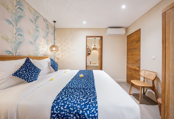 Cozy Balinese bedroom with white king bed, blue floral bedding, frangipani accent, palm-leaf wallpaper, wooden doors, and pendant lighting.