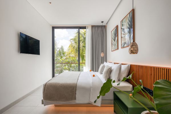 Modern tropical Bali bedroom with king bed, white linens, wooden headboard, green plant, wall-mounted TV, and sliding doors to palm tree views.