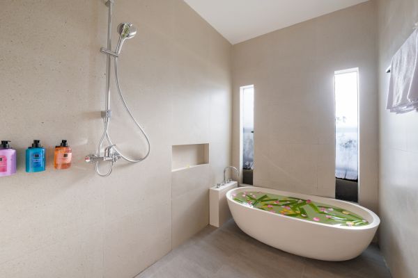 Modern spa bathroom with white freestanding bathtub filled with green water and pink flowers, rainfall shower, and colorful soap bottles. - Bali Villas