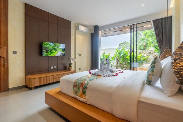 Luxurious Bali villa bedroom with king bed, swan towel decor, wall TV, and sliding doors to lush tropical garden.