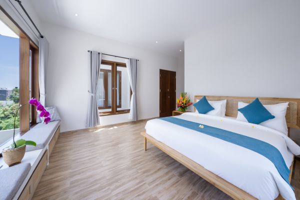 Spacious Balinese-style bedroom with wooden bed, white linens, blue pillows, large window with greenery view, and potted orchids.