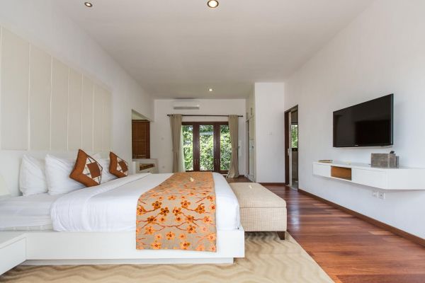 Spacious modern bedroom in a tropical villa, featuring a king bed with orange floral bedding, wall-mounted TV, and sliding glass doors to a lush garden view. - Bali Villas