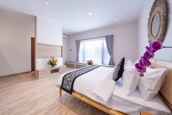 Spacious Balinese bedroom with wooden bed, white linens, purple orchids, tropical flowers, and rattan mirror.