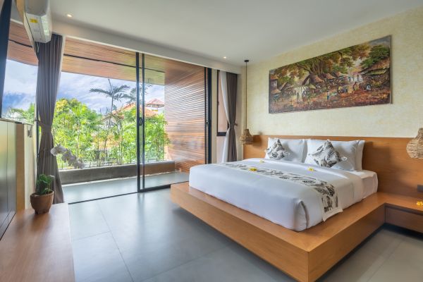 Spacious Balinese bedroom with wooden platform bed, white linens adorned with frangipani, and lush tropical garden view through large sliding glass doors.