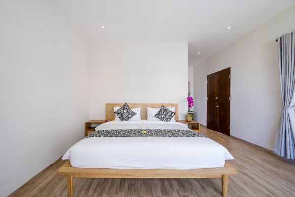 Minimalist Balinese bedroom with white wooden bed, batik pillows, orchid vase, and sheer curtains.