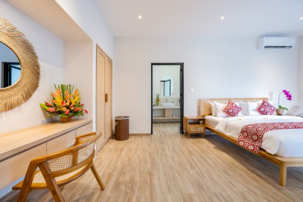 Spacious tropical bedroom with king bed, rattan desk and chair, woven mirror, and colorful floral accents in modern Balinese style.