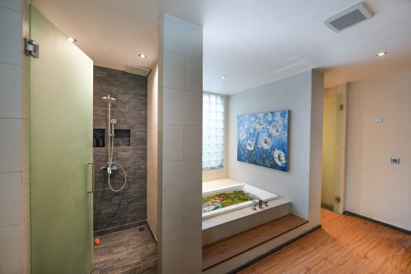 Modern luxury bathroom with green glass shower enclosure, petal-filled soaking tub, and large blue floral wall painting. - Bali Villas