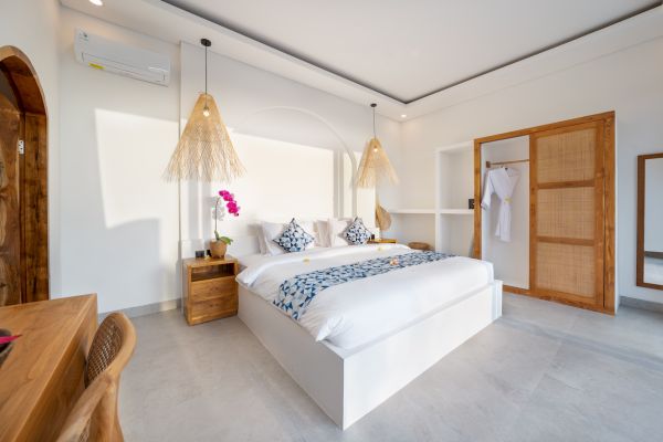 Spacious Balinese bedroom with white king bed, blue-patterned linens, pink orchid vase, wooden nightstands, bamboo pendant lamps, and open wardrobe.