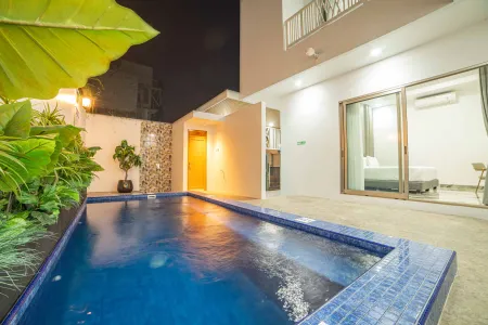 Nighttime view of a modern tropical villa's illuminated blue infinity pool, surrounded by lush greenery and adjacent to a lit bedroom with sliding glass doors.