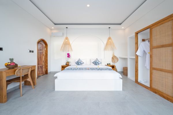 Spacious Balinese-style bedroom with white wooden bed, blue patterned bedding, rattan pendant lights, and tropical wooden furniture.