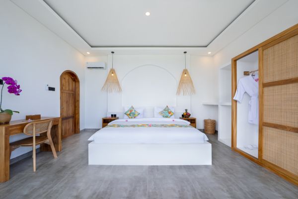 Spacious Balinese-style bedroom with white platform bed, colorful pillows, wooden desk, woven pendant lamps, and potted purple orchids. - Bali Villas