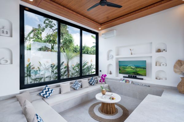 Modern tropical villa living room with white built-in sofa, large sliding glass doors to lush garden, ceiling fan, and wall TV. - Bali Villas