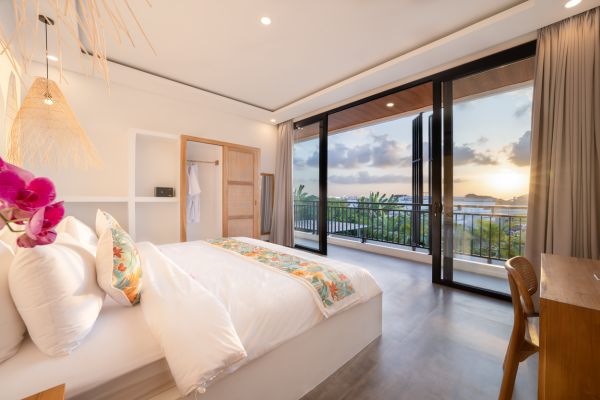 Luxurious Bali bedroom with ocean sunset view from balcony, featuring white bed with floral bedding, pink orchids, and rattan accents.