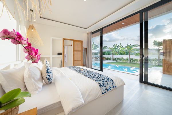 Serene tropical bedroom with white bed, pink orchids, rattan lighting, and sliding glass doors to private infinity pool and palm garden.