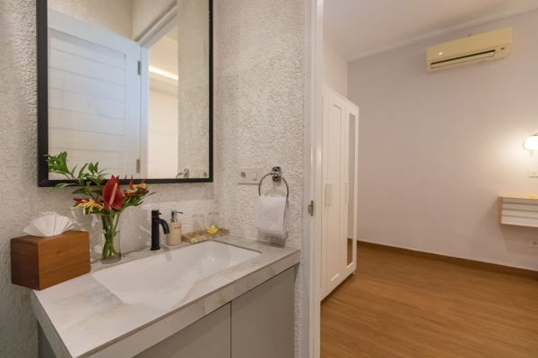 Modern bathroom vanity with white sink, red anthurium flowers in vase, towel holder, and open door to wooden-floored bedroom.