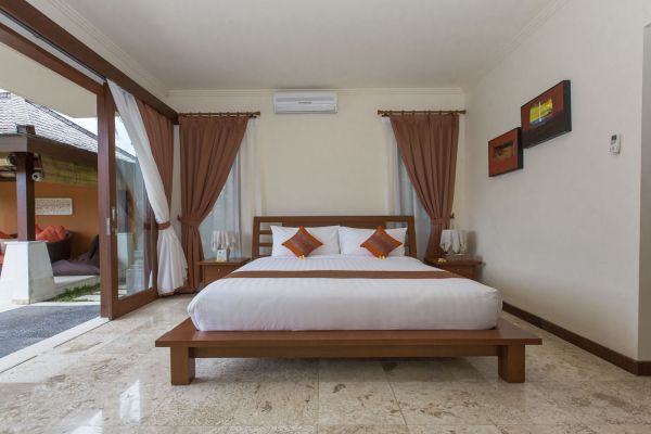 Spacious Balinese villa bedroom with wooden platform bed, white linens, orange pillows, sheer curtains, and sliding doors to tropical patio.