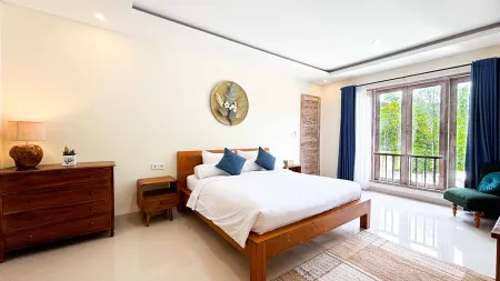 Spacious tropical bedroom with wooden king bed, blue pillows, and large window view of lush greenery.