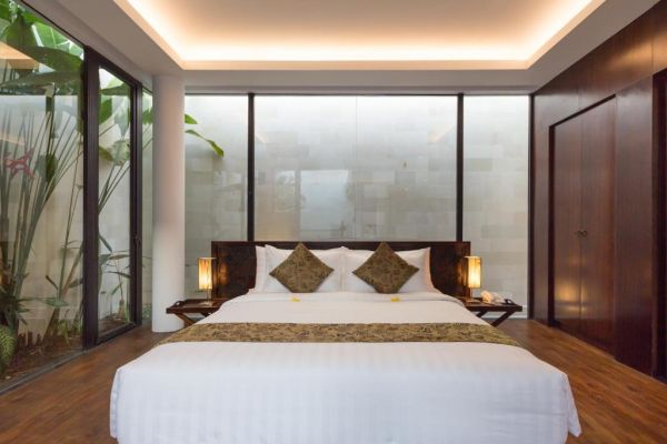 Luxurious king-size bed in a modern Balinese villa bedroom with tropical plants, floor-to-ceiling glass walls, and warm wooden accents.