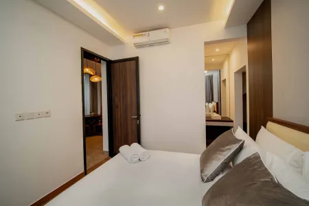Modern minimalist bedroom with a neatly made bed featuring white linens, gray velvet pillows, and folded towels, adjacent to a warmly lit walk-in area and air-conditioned space.
