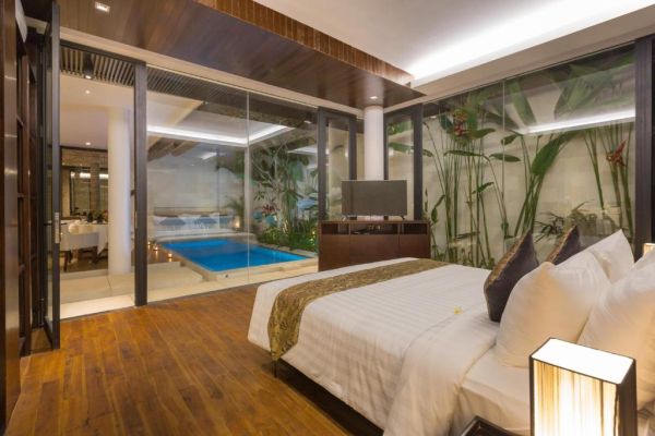 Luxurious tropical bedroom with king bed, modern wooden floors, and large glass doors opening to a private infinity pool amid lush plants.
