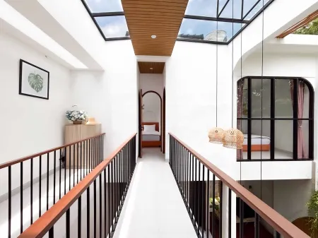 Modern tropical villa interior with glass skylight, elevated walkway, white walls, wooden accents, hanging lanterns, and adjacent bedroom.