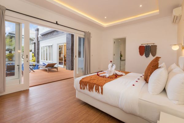 Luxurious Balinese villa bedroom with open sliding doors to a wooden deck and infinity pool, king bed topped with swan towel and rose petals.