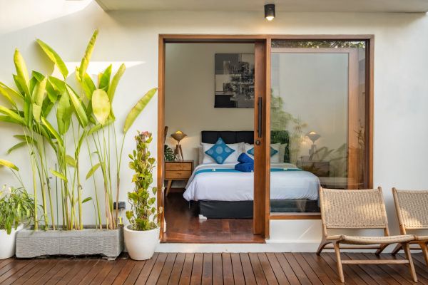 Serene tropical villa bedroom with a king bed in blue linens, open wooden doors to a deck featuring rattan chairs and lush green banana plants.