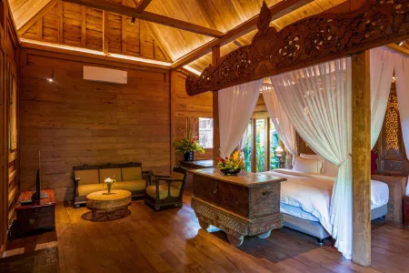 Luxurious Balinese wooden bedroom with ornate canopy bed, teak furniture, tropical plants, and garden-view balcony.
