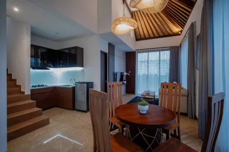 Modern Balinese villa interior featuring a wooden dining table, kitchenette, rattan pendant lights, and large windows with tropical views. - Bali Villas