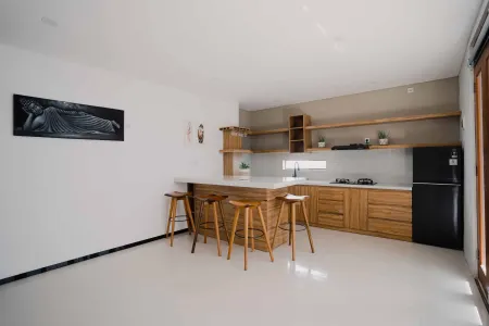 Modern minimalist kitchen with wooden bar counter, stools, cabinets, black refrigerator, and Buddha wall art. - Bali Villas