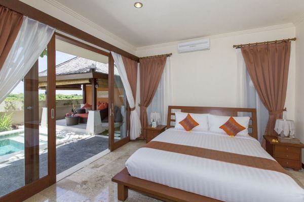 Spacious Balinese villa bedroom with wooden king bed, sheer orange drapes, and sliding doors opening to a private infinity pool.