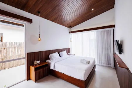 Cozy modern bedroom with wooden ceiling beams, king bed with white linens, wall-mounted TV, and sliding doors to bamboo-fenced balcony.