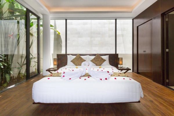 Luxurious tropical bedroom with king-size bed decorated in rose petals and swan-shaped towels, surrounded by floor-to-ceiling windows and lush greenery.