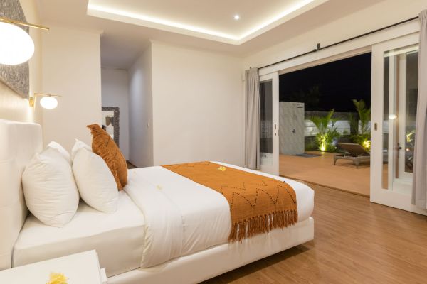 Luxurious modern bedroom with white king bed, orange throw, and open sliding doors to tropical night poolside patio.