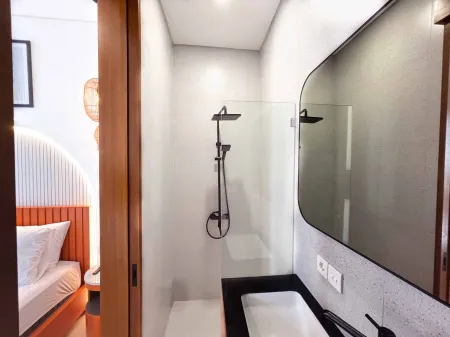 Modern compact bathroom with walk-in glass shower, black vanity sink, and frameless mirror, featuring wooden accents and adjacent bedroom view.