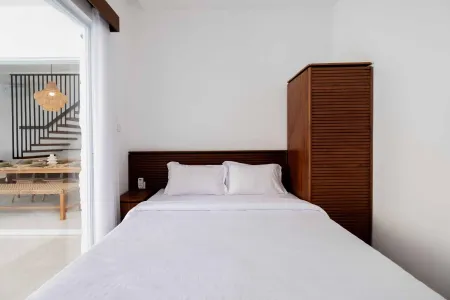 Modern minimalist bedroom with white linen bed, slatted wooden wardrobe, rattan pendant light, and sliding doors opening to a tropical patio dining area.