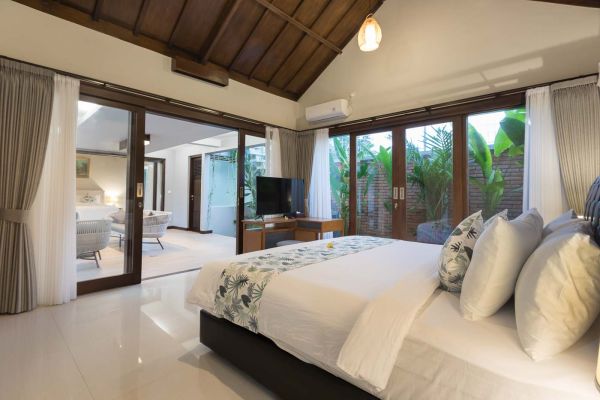 Spacious Balinese-style bedroom with king bed, leaf-patterned duvet, large glass doors to tropical garden, and wooden beamed ceiling.