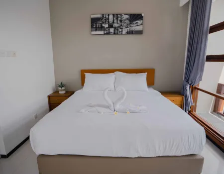 Cozy hotel bedroom with white king bed adorned by two towel swans, wooden headboard, abstract wall art, potted plant on nightstand, and large window with blue curtains.