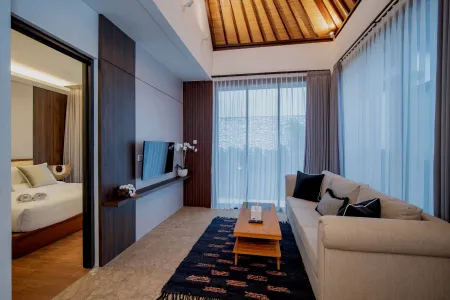 Spacious Balinese-style hotel suite featuring a wooden beamed ceiling, plush beige sofa, low coffee table on a patterned rug, mounted TV, and sheer-curtained windows with natural light. - Bali Villas