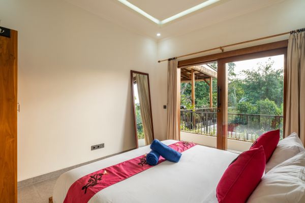 Spacious tropical bedroom with red-accented king bed, full-length mirror, and sliding doors to lush green balcony view.
