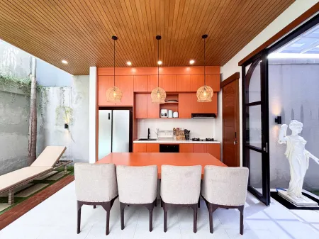 Modern tropical villa kitchen and dining area with orange cabinets, wooden ceiling, pendant lights, and outdoor pool view featuring a statue. - Bali Villas