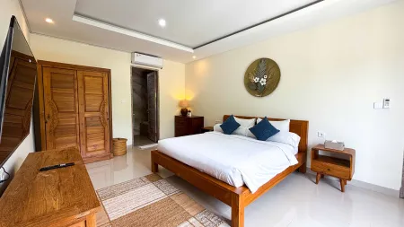Spacious Balinese-style bedroom with wooden king bed, blue pillows, wall-mounted TV, and tropical wall art on light yellow walls.