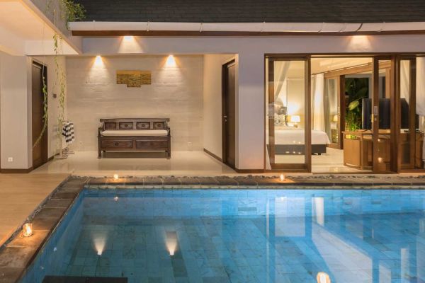 Nighttime view of a candle-lit private pool beside open glass doors to a luxurious bedroom in a Balinese-style villa, with a wooden bench on the terrace.