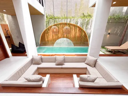 Modern tropical villa interior with turquoise infinity pool, beige L-shaped sofa, lush greenery, and arched bamboo screen. - Bali Villas